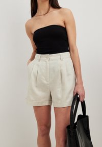 Beige linshorts med veck och rullad fåll, kombinerade med en svart strapless topp. Modellen håller en svart läderhandväska.