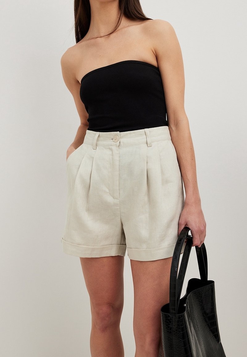 Beige linshorts med veck och rullad fåll, kombinerade med en svart strapless topp. Modellen håller en svart läderhandväska.