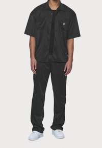Camicia nera a maniche corte con bottoni, dotata di due tasche frontali, abbinata a pantaloni neri e sneakers bianche, con un piccolo logo sulla camicia.