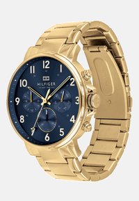 Montre-bracelet en métal doré avec un cadran bleu foncé, des index horaires dorés, trois chronomètres et le logo "Hilfiger EST 1985" sur le cadran.