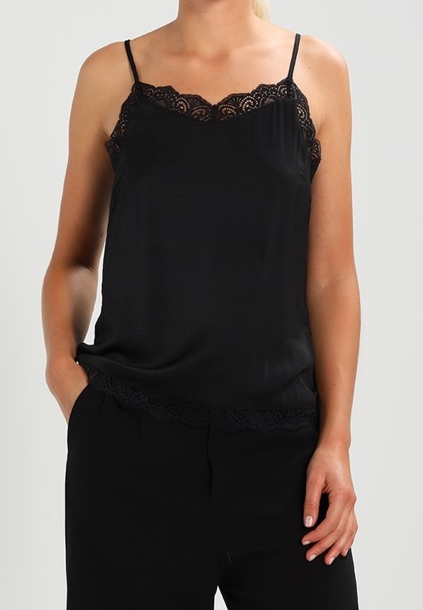 Femme portant un débardeur noir bordé de dentelle et un pantalon noir, debout avec une main dans la poche et une autre reposant sur le côté.