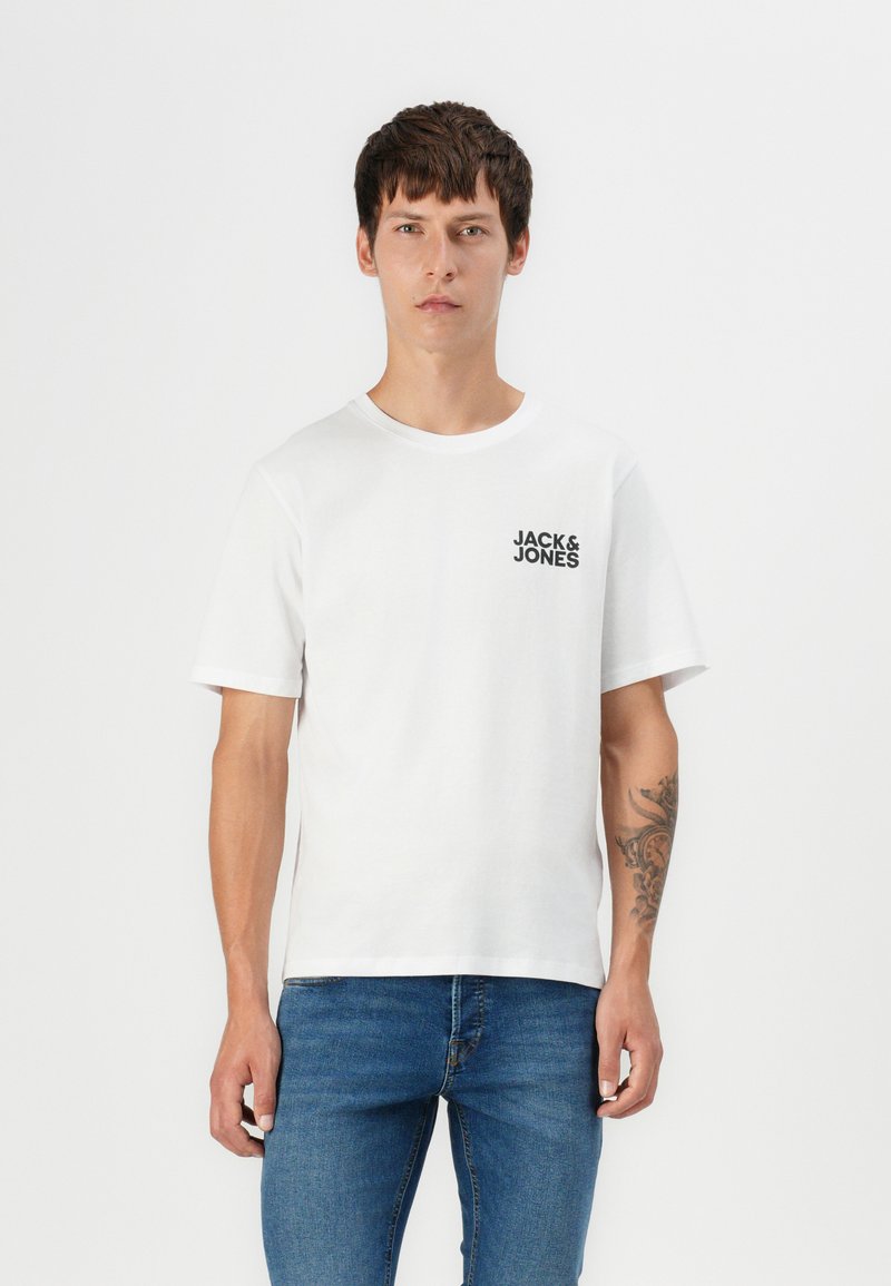 Hvit bomulls T-skjorte med korte ermer, rund halslinje og svart logo på venstre bryst. Kombinert med blåjeans, har modellen en tatovering på armen.