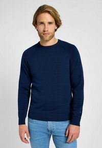 Granatowy sweter z dzianiny z okrągłym dekoltem, ściągaczami na rękawach i u dołu. Charakteryzuje się gładką fakturą i dopasowanym krojem, noszony na jasnoniebieskie dżinsy.