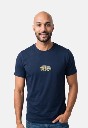 Uomo con barba corta e testa rasata che indossa una maglietta blu navy con un disegno grafico giallo sul petto e jeans blu.