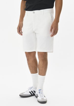 White cotton shorts with a button closure, straight cut, and side pockets, paired with white textured socks and black-striped sneakers.
