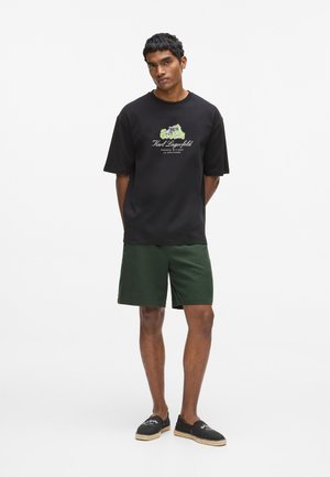 Čierne bavlnené tričko s krátkymi rukávmi a zelenou grafickou potlačou; v páre s tmavozelenými šortkami a čiernymi slip-on topánkami s jútovým lemom.