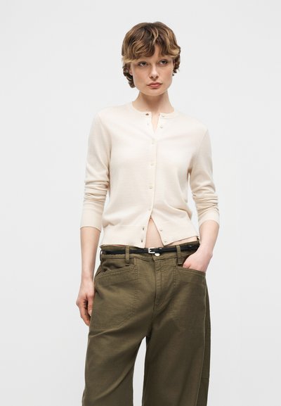 Jeune personne portant un cardigan crème boutonné et un pantalon ample vert olive, les mains dans les poches, debout devant un fond clair uni.