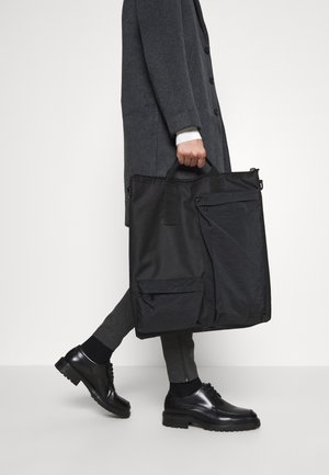 Osoba w ciemnoszarym płaszczu i spodniach, niosąca dużą czarną torbę typu tote z wieloma zamykanymi na zamek kieszeniami, ubrana w czarne eleganckie buty.