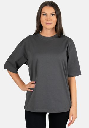 Jonge vrouw met steil bruin haar die een los donkergrijs kort mouw T-shirt draagt, poserend met één hand op haar heup.