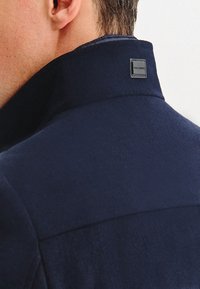 Granatowa kurtka z gładkiego materiału, z wysokim kołnierzem i szarym logo na plecach. Dopasowany krój bez widocznych zapięć.