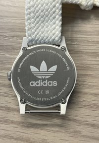 Adidas karóra ezüst rozsdamentes acél hátlappal, fehér szőtt szíjjal és logós részletekkel. Kerek forma sima felülettel.
