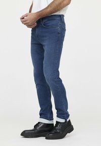 Donkere denim jeans voor mannen met een slim fit, voorzien van een gladde textuur, vijf-pocket ontwerp en opgerolde mouwen boven zwarte schoenen.