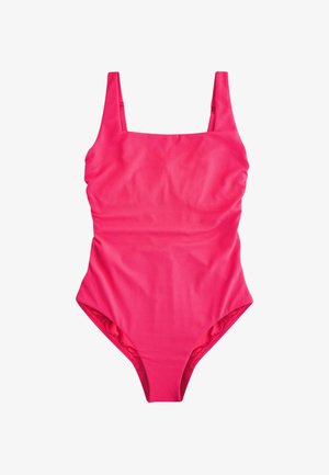 Mujer con un traje de baño rosa de una sola pieza, de pie en una playa de arena con olas del océano y cielo despejado al fondo.