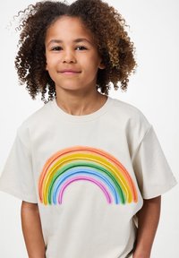 Beżowa koszulka bawełniana z kolorowym, haftowanym wzorem tęczy w kolorach czerwonym, pomarańczowym, żółtym, zielonym, niebieskim i fioletowym. Krótkie rękawy, okrągły dekolt.