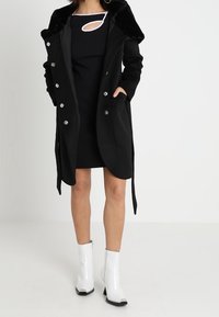 Abrigo negro con cuello de piel, cierre de botones y bolsillos laterales, llevado sobre un vestido negro con escote. La modelo lleva botas blancas hasta el tobillo.