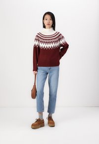 Bordeaux gebreid sweater met een wit en rode geometrische print, hoge kraag, gecombineerd met lichtblauwe jeans en tan platform schoenen. Bruine handtas.