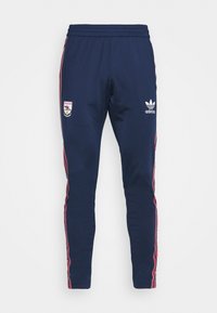 Námořnické sportovní kalhoty s elastickým pasem, logem Adidas a červenými pruhy po stranách. Na levé noze je znak klubu, hladká textura látky.