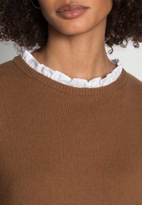 Brązowy sweter z prążkowanej tkaniny, z okrągłym dekoltem. Posiada widoczną białą falbanę pod szyją, co dodaje tekstury i kontrastu.