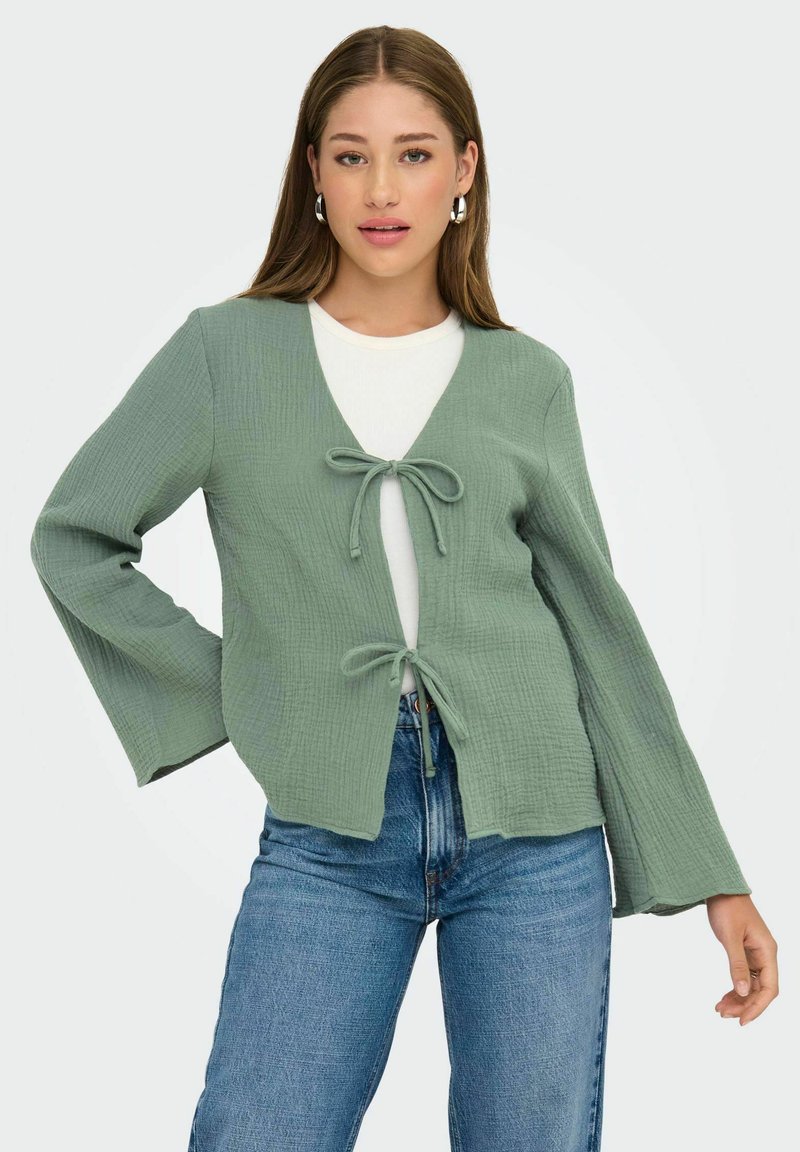 Cárdigan de color verde claro, texturizado, con escote en V, cierre de lazo en el frente y mangas largas acampanadas, usado sobre una blusa blanca y jeans azules.