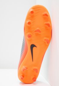 Sole fotbalových kopaček Nike, s jasně oranžovou gumou, texturovaným gripem a šesti kolíky, černým logem a šedým pruhovaným povrchem.