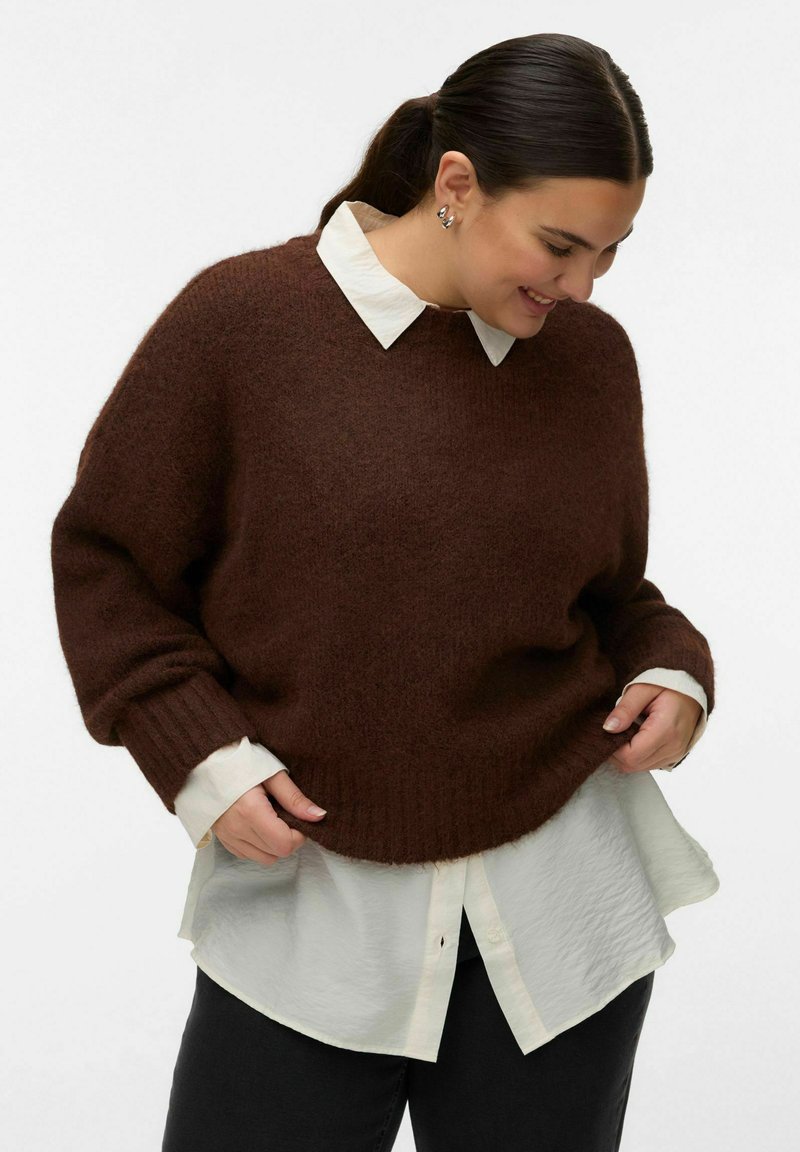 Femme aux cheveux foncés attachés en queue de cheval, souriante, portant un pull marron sur une chemise blanche à boutons et un pantalon noir, sur un fond uni.