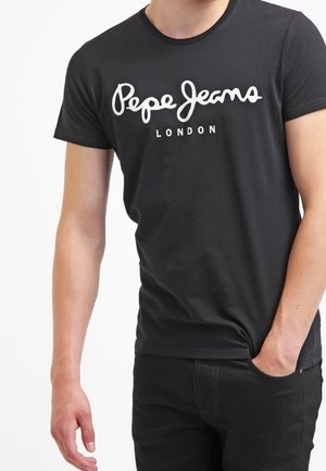 Mies, jolla on musta Pepe Jeans London -t-paita ja mustat housut, toinen käsi taskussa ja toinen rentona sivulla.