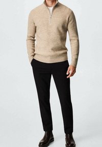Uomo che indossa un maglione beige a zip a quarti, pantaloni neri e stivali marrone scuro, in piedi con una mano in tasca su uno sfondo semplice.