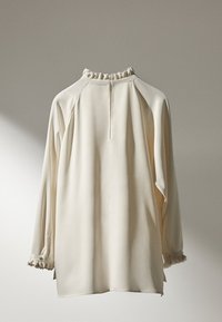 Blouse couleur crème avec un col à volants haut, de longues manches raglan et des poignets boutonnés. Tissu lisse avec un drapé élégant.