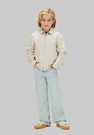 Ungt barn med blondt hår står med hænderne i lommerne iført beige jakke, lyseblå jeans og hvide sneakers med tanfarvet sål.