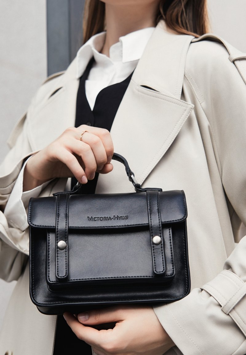 Femme tenant un sac à main en cuir noir Victoria Hyde avec poignée supérieure, portant un trench-coat beige et une chemise blanche avec un gilet noir.
