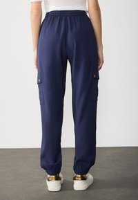 Pantaloni cargo blu navy con cintura elastica, tasche laterali e orli a coste, realizzati in tessuto morbido, abbinati a sneakers bianche e dorate.