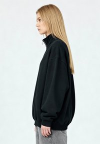 Pull-over en polaire noire avec col montant et fermeture éclair. Comprend des épaules tombantes, des manches longues et des poignets côtelés, créant une silhouette décontractée.