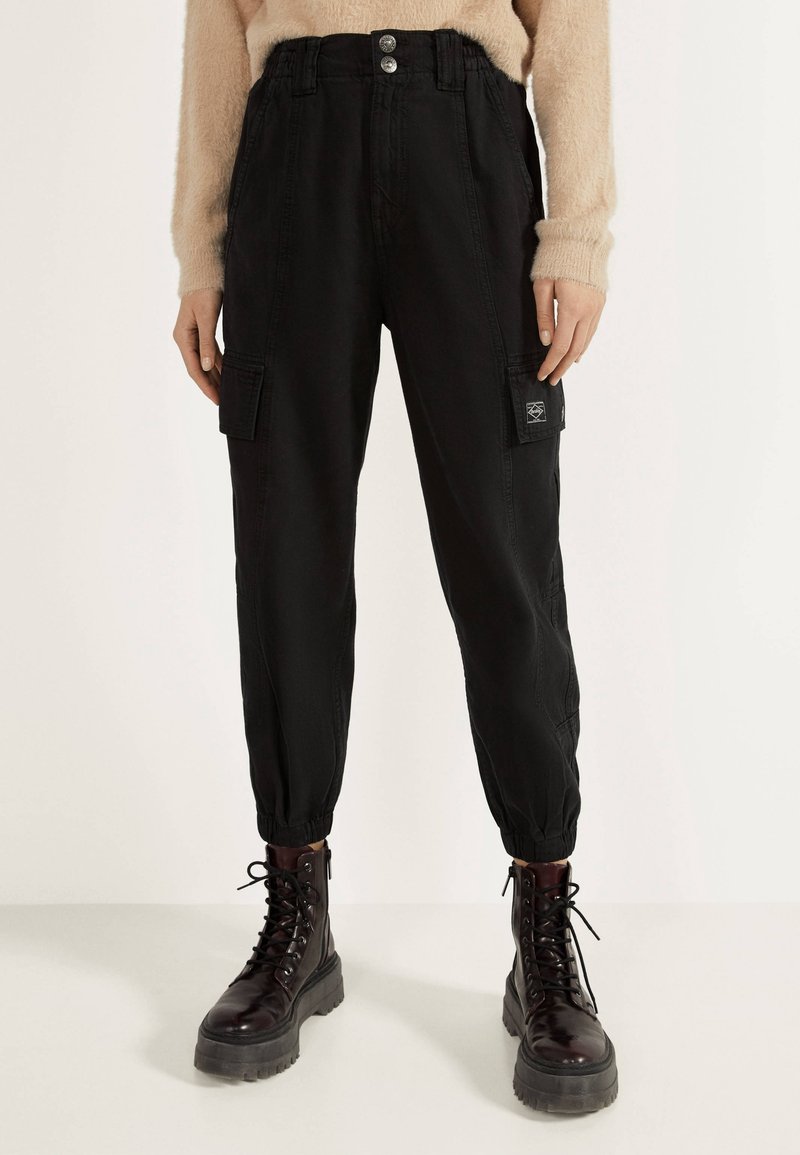 Pantalon cargo noir taille haute avec poignets élastiques, associé à des bottes de combat marron foncé à lacets, sur une personne portant un pull beige duveteux.
