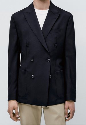 Homme portant un blazer noir croisé à six boutons, poches avant, et un pantalon beige, debout devant un fond uni.