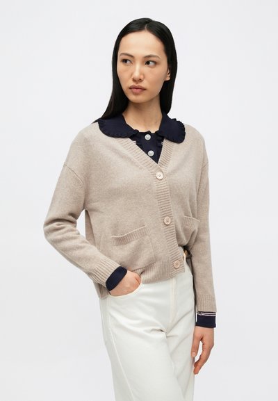 Γυναίκα με μπεζ ζακέτα με κουμπιά πάνω από μπλε πουκάμισο με βολάν στον γιακά και λευκό παντελόνι, όρθια με το ένα χέρι στην τσέπη, κοιτάζοντας προς τα αριστερά.