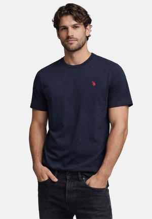 Man with short dark hair wearing a navy blue short-sleeve t-shirt with a small red logo, hands in black jeans pockets, neutral background.