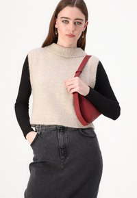 Maglione senza maniche beige lavorato a maglia con colletto alto, indossato sopra una camicia nera a maniche lunghe, abbinato a una gonna in denim nera a vita alta e una borsa rossa.
