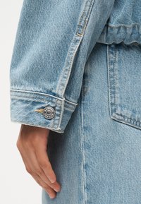 Χέρι που ακουμπάει δίπλα στη μανσέτα ενός ανοιχτού μπλε τζιν μπουφάν με ορατές ραφές και ασημένιο κουμπί στο μανίκι.