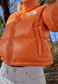 Pomarańczowa kurtka puchowa z wysokim kołnierzem, widocznymi zamkami i kieszeniami bocznymi. Materiał wygląda na gładki i błyszczący w zestawieniu z jasnoszarymi spodniami.