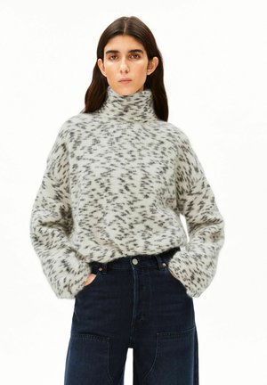 Femme aux longs cheveux foncés portant un pull duveteux à col montant, gris et noir à motif, et un jean taille haute foncé, les mains dans les poches.