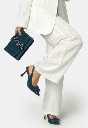 Mujer con traje blanco levanta la pierna, lleva zapatos de tacón alto slingback color verde azulado con hebillas de piedras preciosas, sosteniendo un bolso de mano a juego con cierre de piedras preciosas.