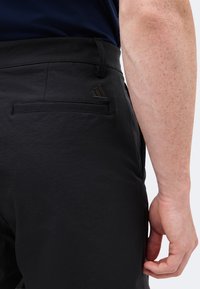 Homme portant un pantalon noir avec une poche arrière et un logo noir brodé, debout avec son bras droit détendu le long du corps.
