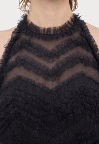 Haut dos nu noir à volants avec tissu transparent et bordure texturée en zigzag autour du décolleté et de la poitrine sur une personne.