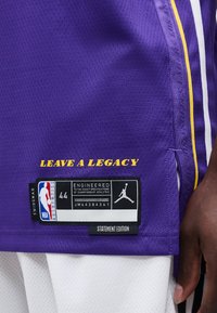 Lilla basketballtrøye med gul tekst "LEAVE A LEGACY", NBA- og Jordan-merkelapper, båret av en person med mørk hud og hvite shorts.