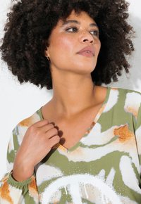 Maglietta a maniche lunghe con scollo a V in verde, decorata con un leggero motivo astratto e un segno di pace bianco. Presenta polsini arricciati e una texture morbida.