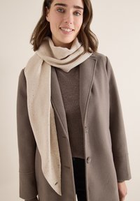 Écharpe en tricot beige avec de petits points brun doré, superposée à un pull brun et un manteau gris. Texture douce et forme longue et drapée.