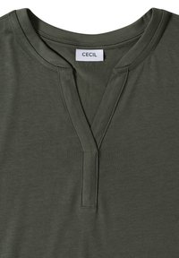 Chemise vert olive avec un col rond fendu et une étiquette cousue portant l'inscription "CECIL" au niveau du col.