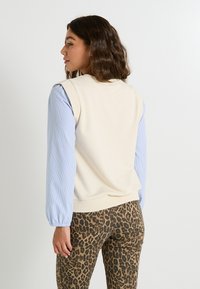 Top sin mangas de color crema con cuello redondo, combinado con una camisa de rayas azul clara de manga larga; usado con pantalones de leopardo marrón. Textura suave, ajuste casual.
