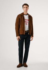 Giacca in suede marrone con chiusura a zip, abbinata a una camicia bianca con testo rosso, jeans blu scuro e stivali alla caviglia in suede marrone.