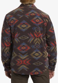 Langsleeve shirt van donkerbruin materiaal met levendige, multikleurige geometrische patronen, waaronder diamanten en pijlen, met een zachte textuur.